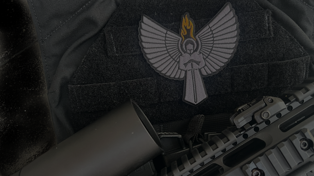 A tactical embroidered patch featuring a stylized winged emblem with a flame and central symbol, shown on a black vest next to a rifle in a dark, moody setting.